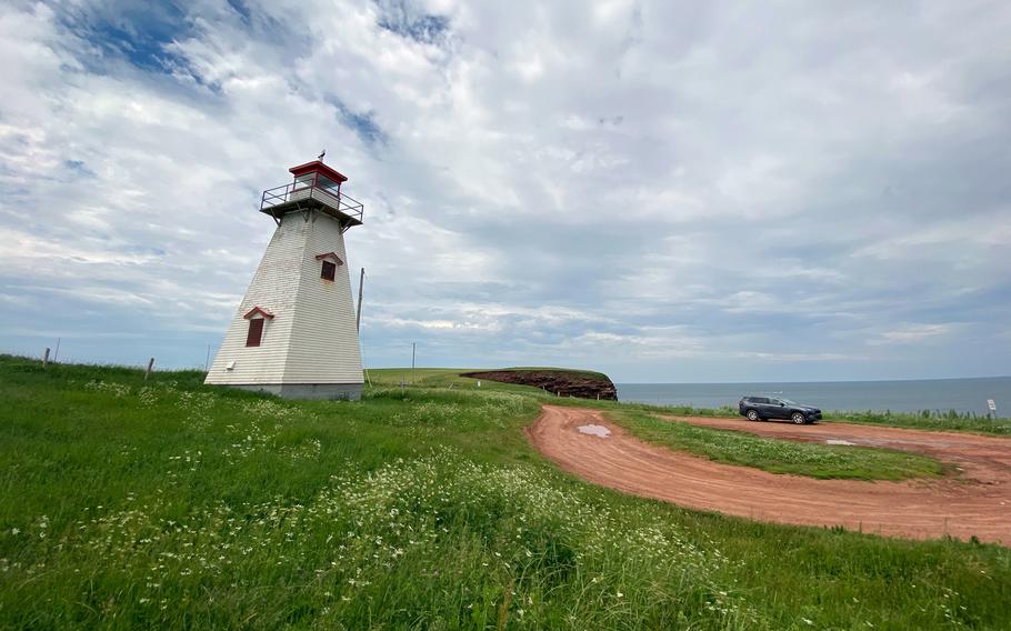 A lighthouse stands on a grassy dune with a dirt road winding next to it and the horizon above the ocean in the background.