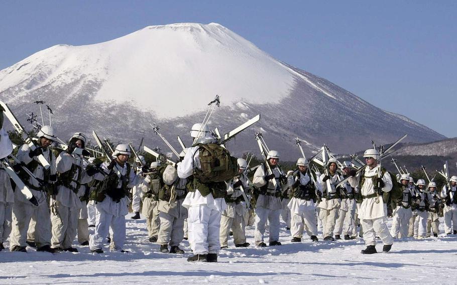 Service members in white protective clothes stand in a snowy area with a snow-capped volcano in the background.