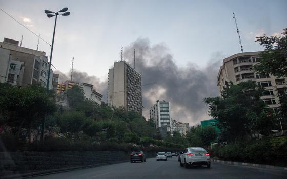 Smoke rises over multi-story buildings in Tehran, Iran, as cars drive on a road in the foreground.
