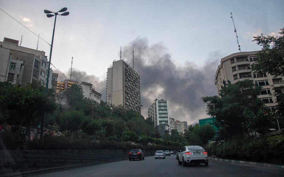 Smoke rises over multi-story buildings in Tehran, Iran, as cars drive on a road in the foreground.