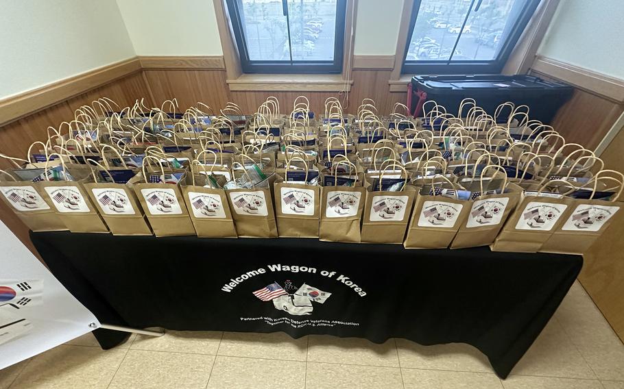 Several dozen brown paper grocery bags with white signs reading “Welcome Wagon of Korea” and pictures of U.S. and South Korean flags rest on a table covered by a black tablecloth with the same “Welcome Wagon of Korea” emblem and flag pictures.