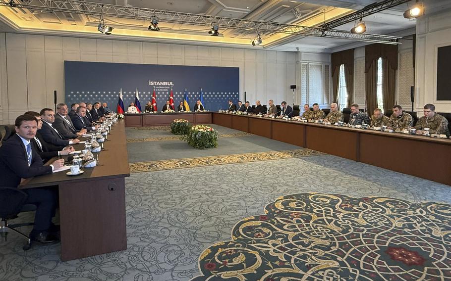 Government officials sit at a large U-shaped table in a conference room with Russian and Ukrainian flags in the background.