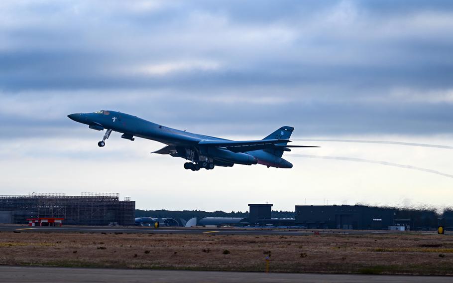 A U.S. Air Force strategic bomber takes off from an airfield under a cloudy sky. The aircraft is slightly tilted upward as it ascends, with landing gear still extended and heat distortion visible from its engines. In the background, airport infrastructure, including hangars and a control tower, is seen on the tarmac surrounded by grassy terrain.