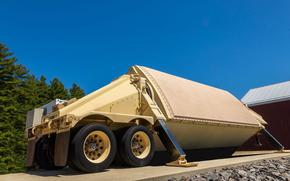 A large, tan-colored military radar system is mounted on a wheeled trailer, positioned at an angle on a concrete platform. The radar is housed in a protective, angular enclosure, with stabilizing legs extended to the ground. 