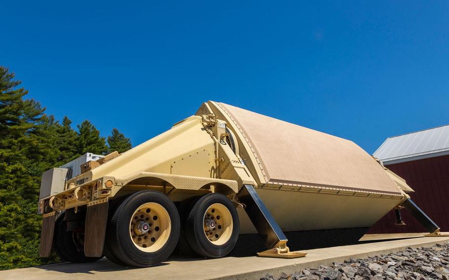 A large, tan-colored military radar system is mounted on a wheeled trailer, positioned at an angle on a concrete platform. The radar is housed in a protective, angular enclosure, with stabilizing legs extended to the ground. 