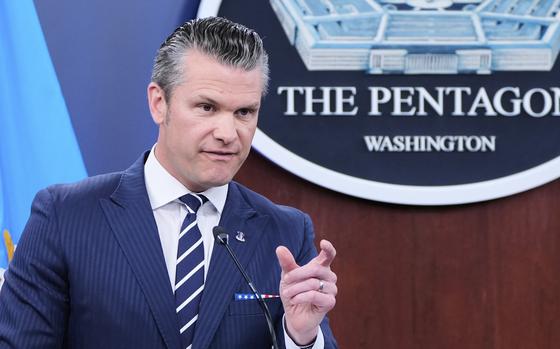 Pete Hegseth, wearing a dark suit, speaks into a microphone and gestures with his left hand as he stands in front of a large seal of the Pentagon on the wall behind him.
