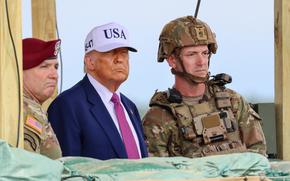 President Donald Trump, wearing a dark suit and red tie while wearing a white “USA” cap,  stands alongside military personnel in combat uniforms and tactical gear. Wooden structures and sandbags are visible in the foreground. 