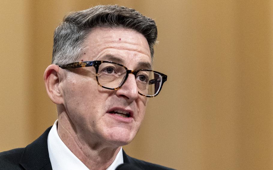 Close-up view of a man in dark rimmed glasses and a dark suit speaking into a microphone in a congressional hearing room.