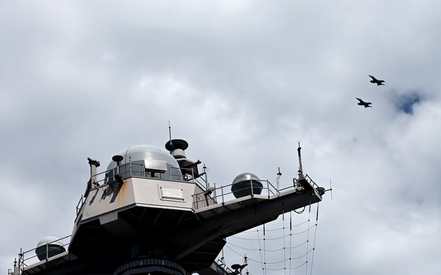 Fighter jets are seen in the sky from the vantage point of an aircraft carrier, with a watch or control tower taking up most of the space.