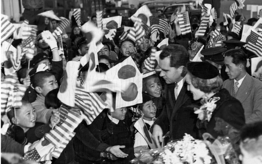 A black-and-white, 1950s-era image of Richard Nixon and his wife before a large group of Japanese children waving U.S. and Japan flags.