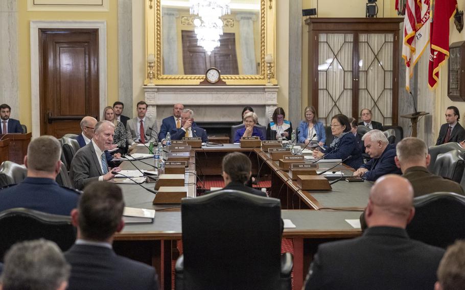 Senators in suits sit around a large rectangular table in a congressional hearing room, with a large mirror above a mantle in the background.