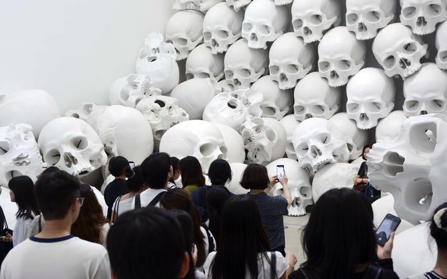 A crowd of spectators use phones to takes photos of a large sculpture composed of stacked human skulls.