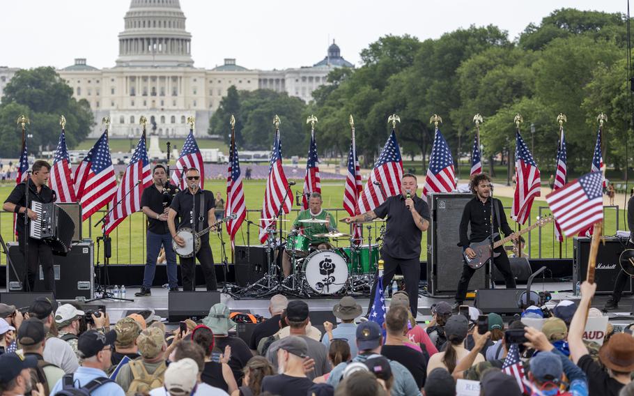A band performs on a stage adorned with U.S. flags on the National Mall, with the U.S. Capitol building in the background.