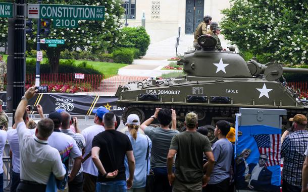 Spectators watch and take pictures of a tank driving down a street.