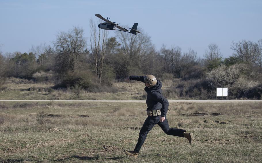 A person in military tactical gear and helmet running across an open field while a military drone flies overhead in the background. 