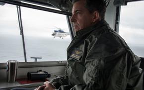 A military officer in a green flight suit sits inside an aircraft carrier gazing out through large windows. Through the windows, another military helicopter can be seen flying in the distance over water. 