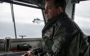 A military officer in a green flight suit sits inside an aircraft carrier gazing out through large windows. Through the windows, another military helicopter can be seen flying in the distance over water. 