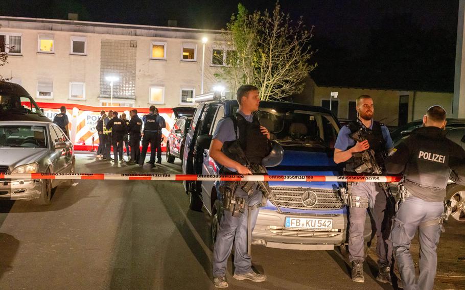 Police officers stand guard at night behind a cordoned-off area in front of a residential building, with several police vehicles and a group of officers gathered near a red and white police barrier. The scene is illuminated by streetlights and vehicle lights.