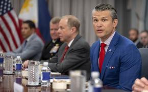 Pete Hegseth in a blue suit and red tie speaking while seated at a conference table.