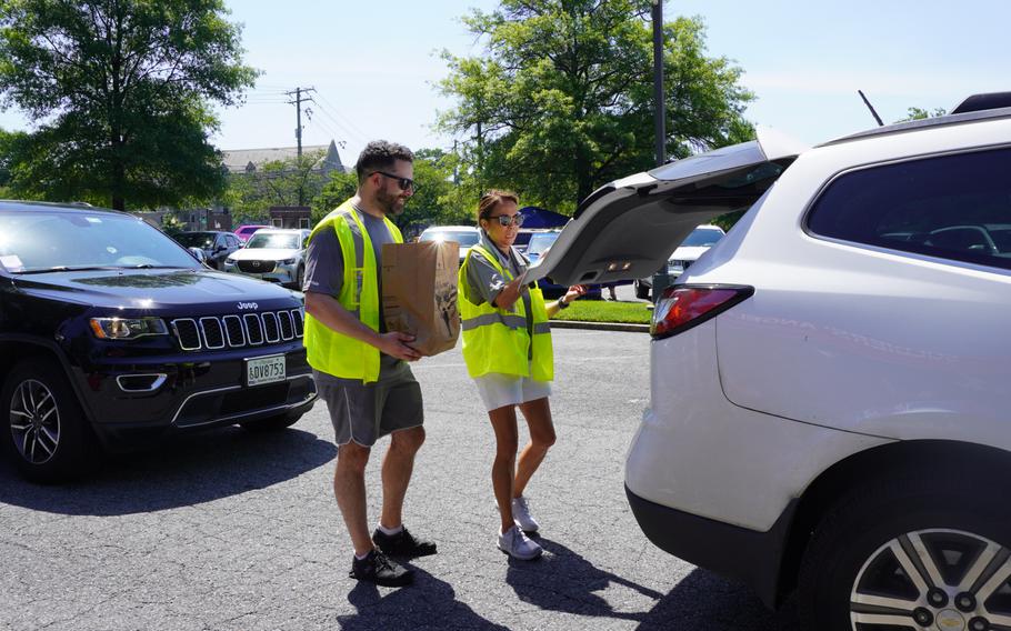A woman opens the trunk of a car for a man holding a paper bag