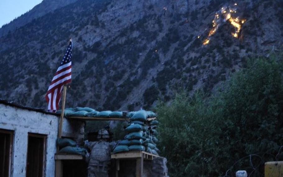 Adam Holroyd looks on as ordnance explodes on a hillside overlooking the Afghan village of Barg-e Matal in the northeastern province of Nuristan in the summer of 2009. An outbuilding that housed hundreds of rocket-propelled grenades and other ammunition, which was later hit by an RPG during an attack, is on the left.