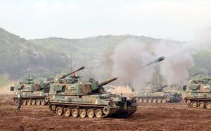 Self-propelled howitzers fire missiles during a drill at a training ground in South Korea, with smoke billowing from their barrels and mountains visible in the background.