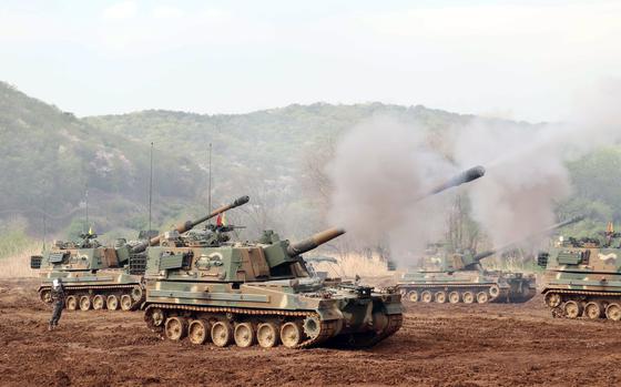 Self-propelled howitzers fire missiles during a drill at a training ground in South Korea, with smoke billowing from their barrels and mountains visible in the background.