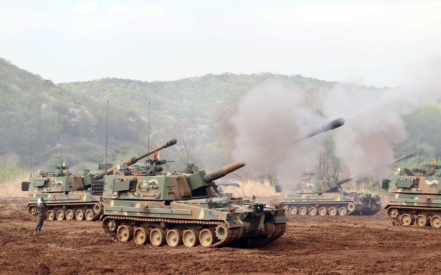 Self-propelled howitzers fire missiles during a drill at a training ground in South Korea, with smoke billowing from their barrels and mountains visible in the background.