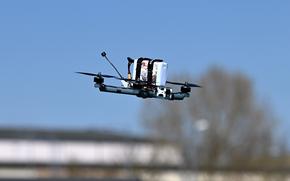 A military drone in flight against a clear blue sky, featuring multiple rotors and various equipment attachments including what appears to be cameras or sensors.