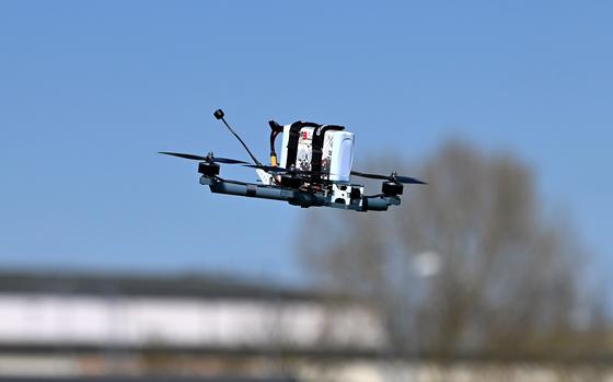 A military drone in flight against a clear blue sky, featuring multiple rotors and various equipment attachments including what appears to be cameras or sensors.