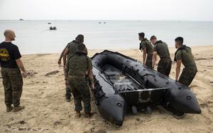 Military personnel in camouflage uniforms carry a large black inflatable boat on a sandy beach, with calm waters and several small boats visible in the background.