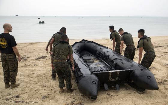 Military personnel in camouflage uniforms carry a large black inflatable boat on a sandy beach, with calm waters and several small boats visible in the background.