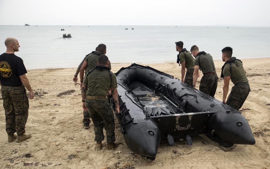 Military personnel in camouflage uniforms carry a large black inflatable boat on a sandy beach, with calm waters and several small boats visible in the background.