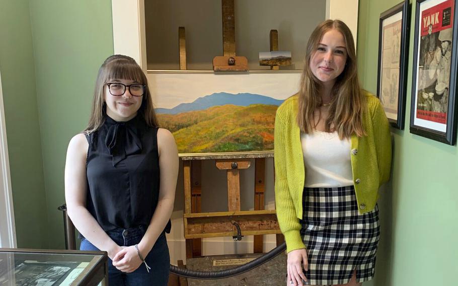 Interning curators Sydney Ball, left, and Lilly Underwood, right, pose in front of Philip Moose’s painting of the Blue Ridge Mountains on July 28, 2022, at the Hickory History Center, in Hickory, N.C. 