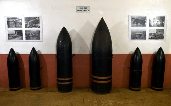 Display of various sizes of dark military artillery shells and munitions arranged against a wall in a museum, with historical photographs visible in the background.