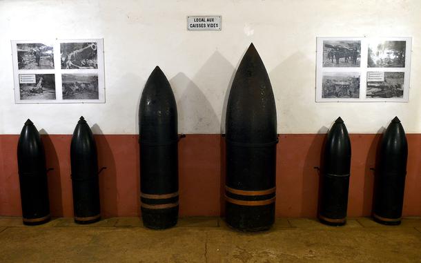 Display of various sizes of dark military artillery shells and munitions arranged against a wall in a museum, with historical photographs visible in the background.