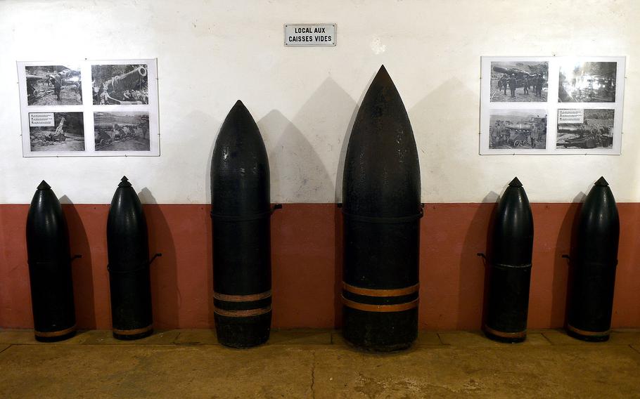 Display of various sizes of dark military artillery shells and munitions arranged against a wall in a museum, with historical photographs visible in the background.