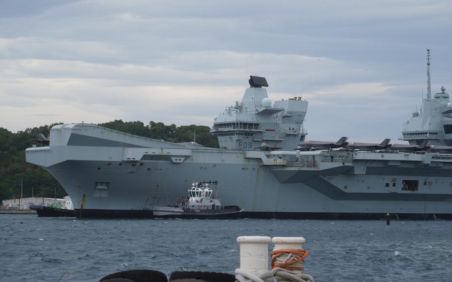 A Royal Navy aircraft carrier is viewed from the water with multiple F-35 fighter jets visible on the flight deck. A small tugboat is positioned alongside the massive grey warship, with forested coastline visible in the background. 