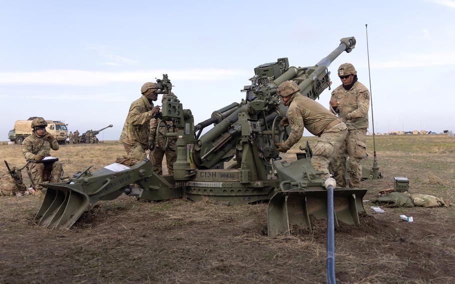 Military personnel in camouflage uniforms operate a green artillery piece in an open field. 