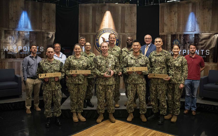 Navy Reserve Lt. Cdr. Jonathan Calhoun, center, holds the i3 Waypoints trophy after Vice Adm. John Mustin, standing behind him, announced Calhoun’s “Leveraging Mobile Technology to Streamline Mobilization” as the winning entry of the inaugural i3 Waypoints, a new invention competition that allows reservists of all ranks to pitch ideas to top brass. Calhoun is surrounded by the other final presenters, the finalist panel and production staff.