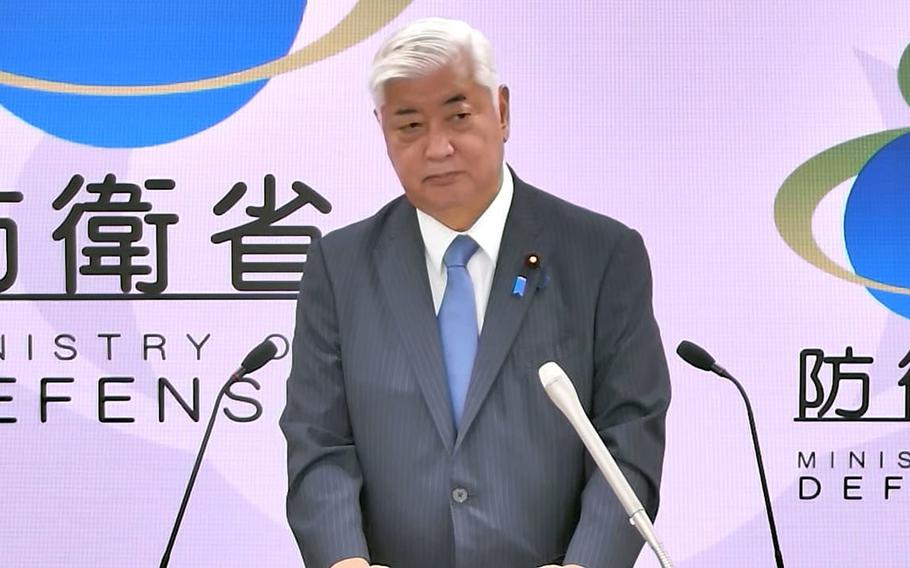 A man in a gray suit and light blue tie stands at a podium with two microphones during a press conference. Behind him is a backdrop featuring the Japanese Ministry of Defense logo along with Japanese characters and “MINISTRY OF DEFENSE” text in both Japanese and English. The background has a soft purple and white gradient design.