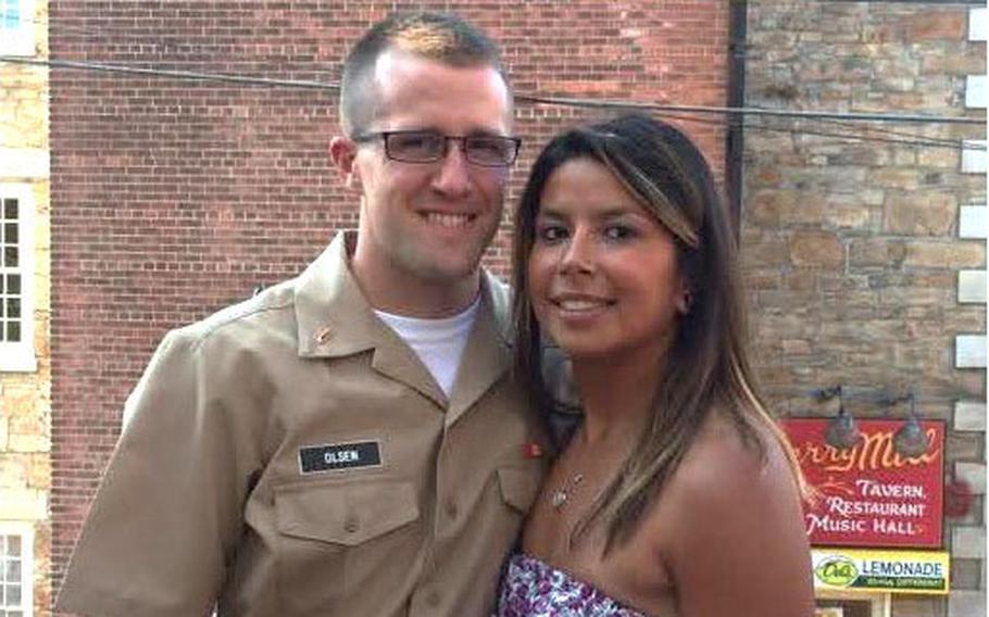 A man in a U.S. Navy officer service khaki uniform and glasses stands next to a woman in a strapless floral dress, both smiling on an outdoor patio with a brick building in the background.