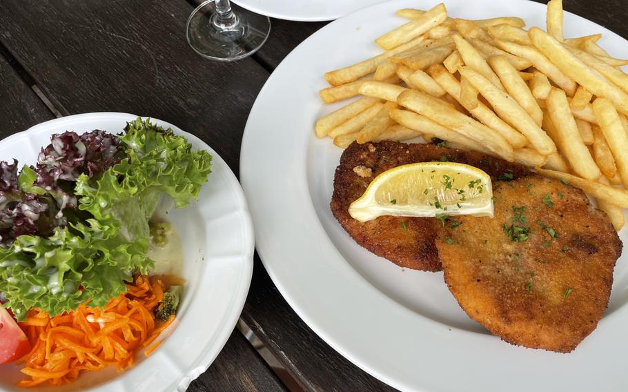 A golden-brown breaded schnitzel served with french fries on a white plate, garnished with a lemon wedge and fresh parsley. A side salad with mixed greens and shredded carrots is visible on a separate plate.