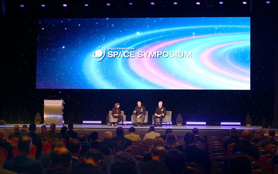 Three people seated on a stage before an audience, underneath a large screen that reads “Space Foundation/SPACE SYMPOSIUM.”