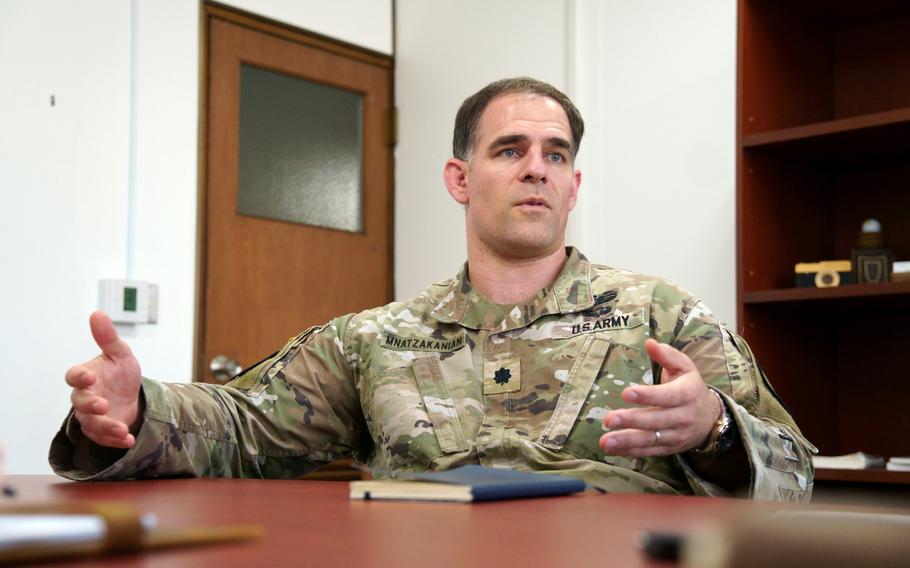 A U.S. Army officer in desert camouflage uniform speaks while seated at a wooden conference table, gesturing with both hands outstretched. 