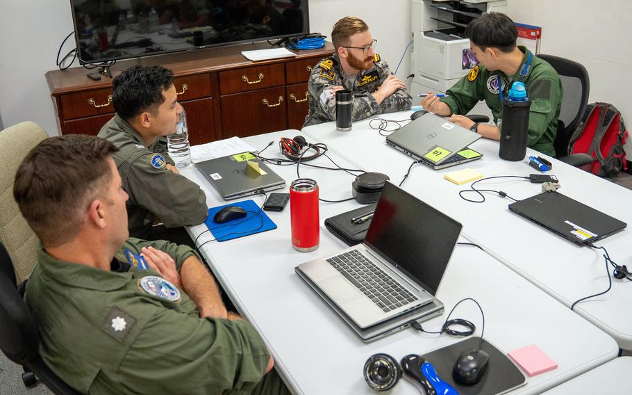 Four military personnel in flight suits work together around a white conference table covered with laptops, sticky notes, water bottles and various electronic equipment. A large monitor and wooden credenza are visible in the background of the office space.