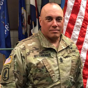 A man in a camouflage Army uniform poses in front of a U.S. flag.
