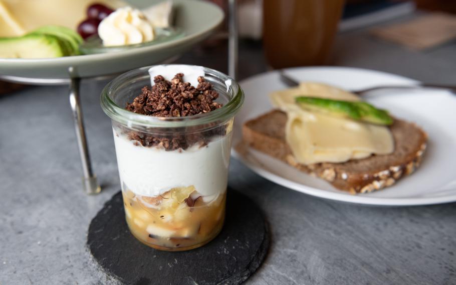 A small pot of chocolate muesli with organic yogurt and fruit rests on a table near two breakfast platters.