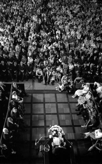 A vertical, aerial and black and white photo of a crowd of people sitting and watching a man speak at a podium.