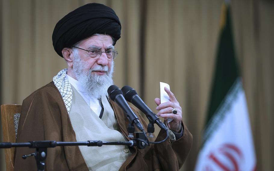 Iran’s supreme leader speaks in front of two microphones while raising a hand.